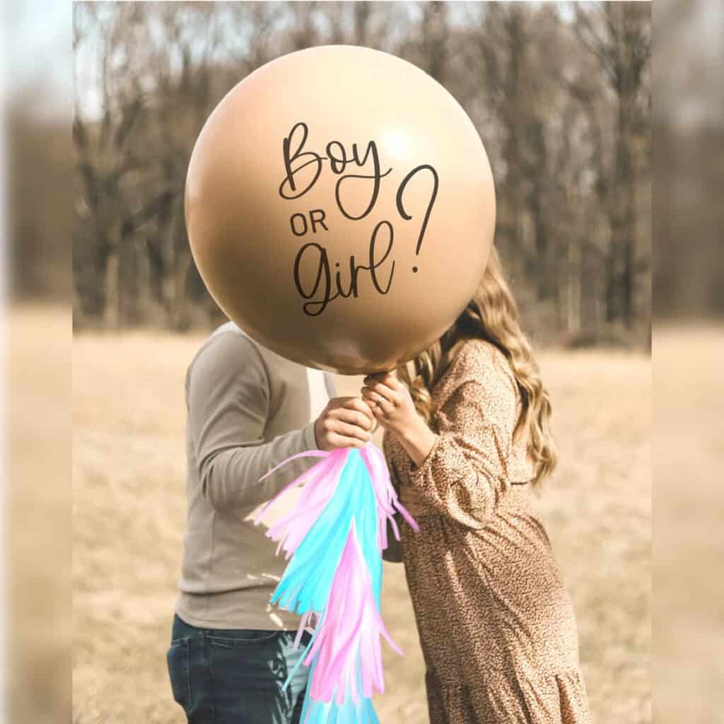 Two people stand outdoors holding a large balloon with Boy or Girl? written on it, showcasing one of the delightful gender reveal ideas. The balloon is adorned with pink and blue tassels in an open field, with trees softly dotting the background.