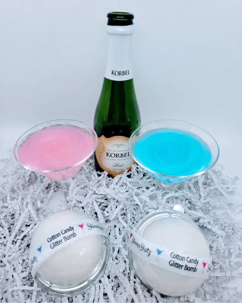 Amid shredded paper, a small bottle of Korbel champagne complements two martini glasses containing pink and blue concoctions—ideal for unique gender reveal ideas. Nearby, cotton candy glitter bomb ornaments add a whimsical touch to the celebratory scene.