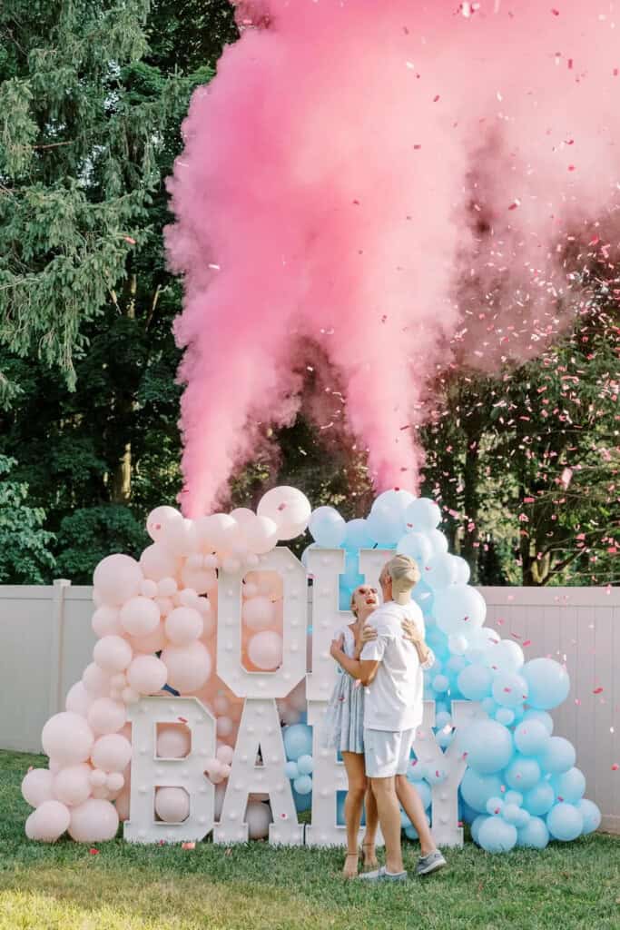 A couple embraces outdoors near a display featuring white, pink, and blue balloons with the words OH BABY. Pink smoke and confetti burst above them in a joyful gender reveal idea. A backdrop of trees and a white fence adds to the celebrations charm.