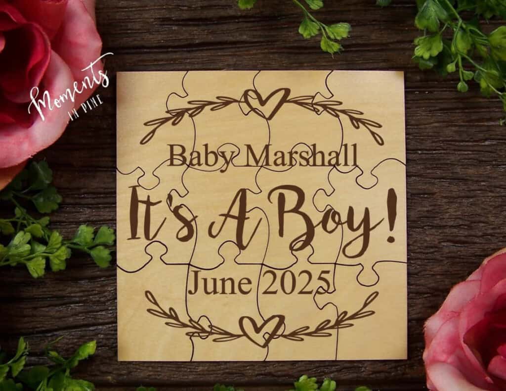 A wooden puzzle, perfect for gender reveal ideas, announces Baby Marshall, Its A Boy! June 2025 with decorative hearts and foliage. Placed on a wooden surface, its surrounded by green leaves and pink flowers.