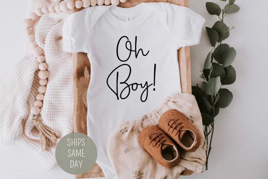 A flat lay image features a white baby onesie with the text Oh Boy! in black, perfect for gender reveal ideas. Its surrounded by knitted fabric, a string of wooden beads, eucalyptus leaves, brown baby shoes, and beige patterned shorts. A sticker reads SHIPS SAME DAY.