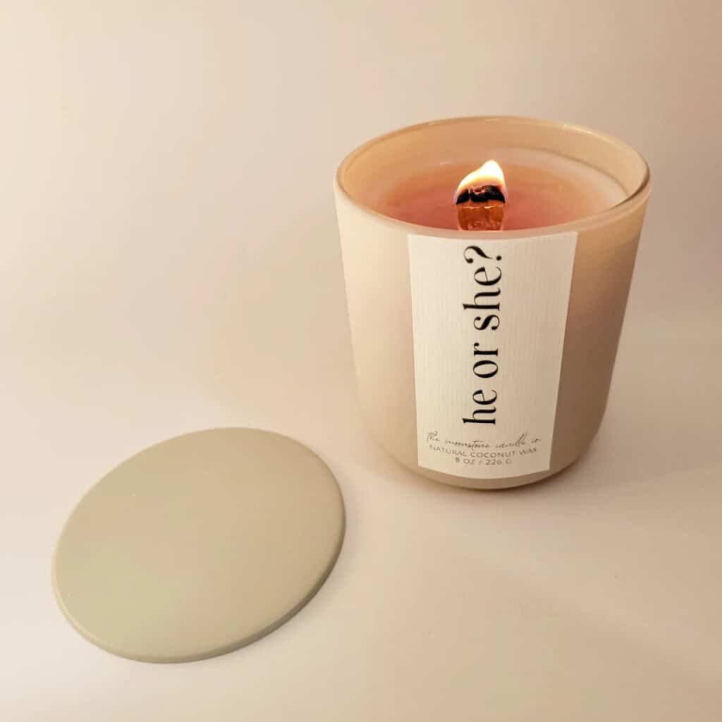 A lit candle with a wooden wick stands on a surface, adding warmth to your gender reveal ideas. The candle is in a beige container with a label that reads he or she? and is complemented by a matching lid beside it. Inside, the wax glows light pink, setting the perfect ambiance for the occasion.