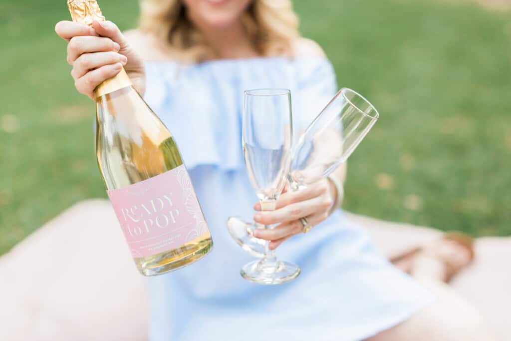 A person in a light blue dress holds a Ready to Pop champagne bottle and two empty flutes, perfect for creative gender reveal ideas. The scene unfolds outdoors on a lush grassy surface, inviting anticipation and celebration.