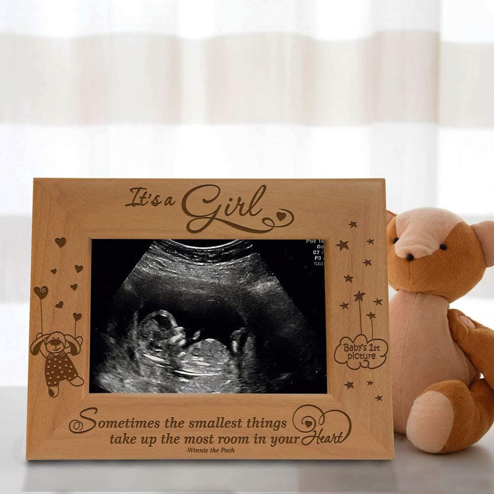 A tan picture frame containing a sonogram sits beside an animal plush with striped white and gray curtains in the background. The top of the frame reads "It's a Girl" with a heart. The bottom reads "Sometimes the smallest things take up the most room in your Hearts -- Winnie the Pooh." The left side of the frame features an image of a bunny in overalls holding heart-shaped balloons, and the right side features the text "Baby's 1st pictures" in a cloud surrounded by stars.