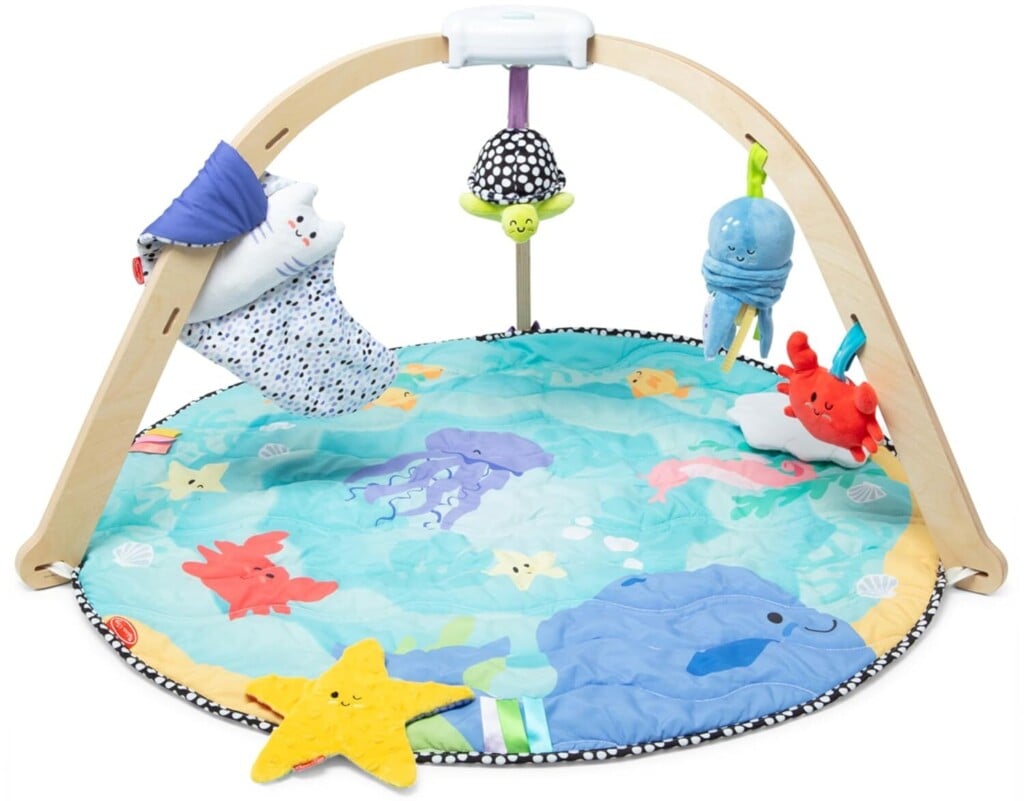 A colorful baby play mat, part of our Baby Products for 2025 collection, features a wooden arch frame with hanging toys, including a plush white bear and a blue octopus. The round mat dazzles with sea-themed designs like starfish and seashells on a serene blue background.