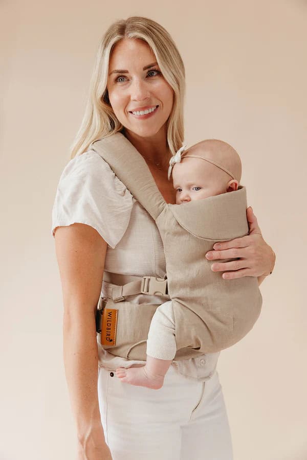 A person with long blonde hair, sporting a white blouse and light pants, cradles a baby in a beige baby carrier. The babys headband complements their cozy position against the persons chest. This scene captures the essence of Baby Products for 2025 against a plain, light background.