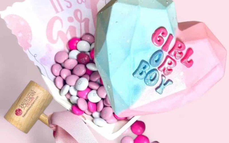 A pink and blue heart-shaped confection reading GIRL OR BOY crowns a small container of pink and white candy. A pink and brown mallet waits nearby, alongside a card proclaiming Its a Girl, making it one of the sweetest gender reveal ideas.