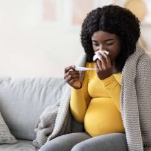 A pregnant woman sits on a gray couch, wrapped in a knitted blanket, feeling the effects of getting sick while pregnant. Clad in a yellow shirt and gray pants, she checks her temperature with a thermometer and reaches for home remedies for cold during pregnancy.