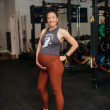A pregnant woman stands in a gym wearing a tank top and leggings. She rests one hand on her belly and smiles. The gym is equipped with various fitness equipment, including weights and resistance bands, in the background.