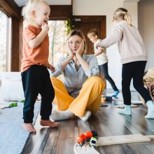 A 21st century mom sits on the floor, hands on her cheeks, looking contemplative. Around her, three children joyfully play with toy trains and tracks. A cat ambles by in the room filled with scattered toys and plants, lit by the light from large windows.