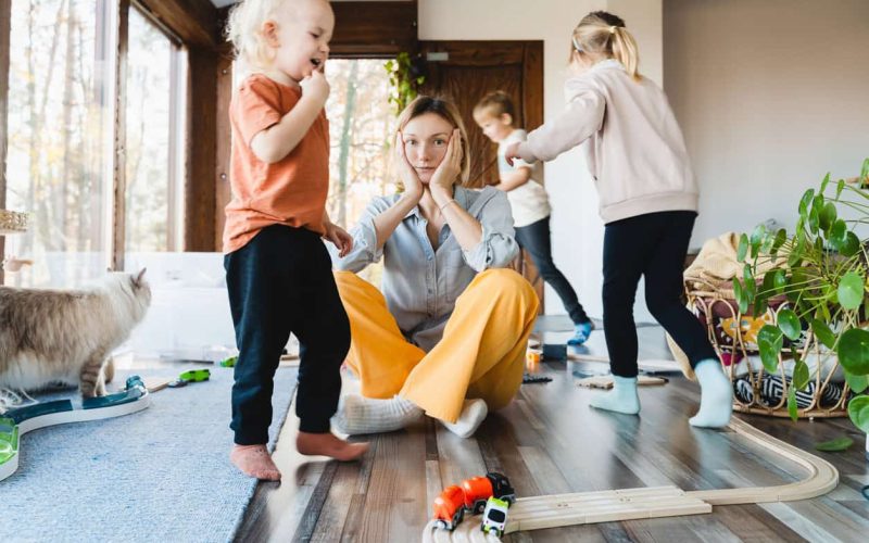 A 21st century mom sits on the floor, hands on her cheeks, looking contemplative. Around her, three children joyfully play with toy trains and tracks. A cat ambles by in the room filled with scattered toys and plants, lit by the light from large windows.