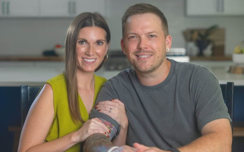 Whitney and Brandon Cawood sit closely together at a table. She has her hand resting on his arm. They are both smiling warmly, surrounded by a kitchens white cabinets, creating an atmosphere as inviting and vibrant.